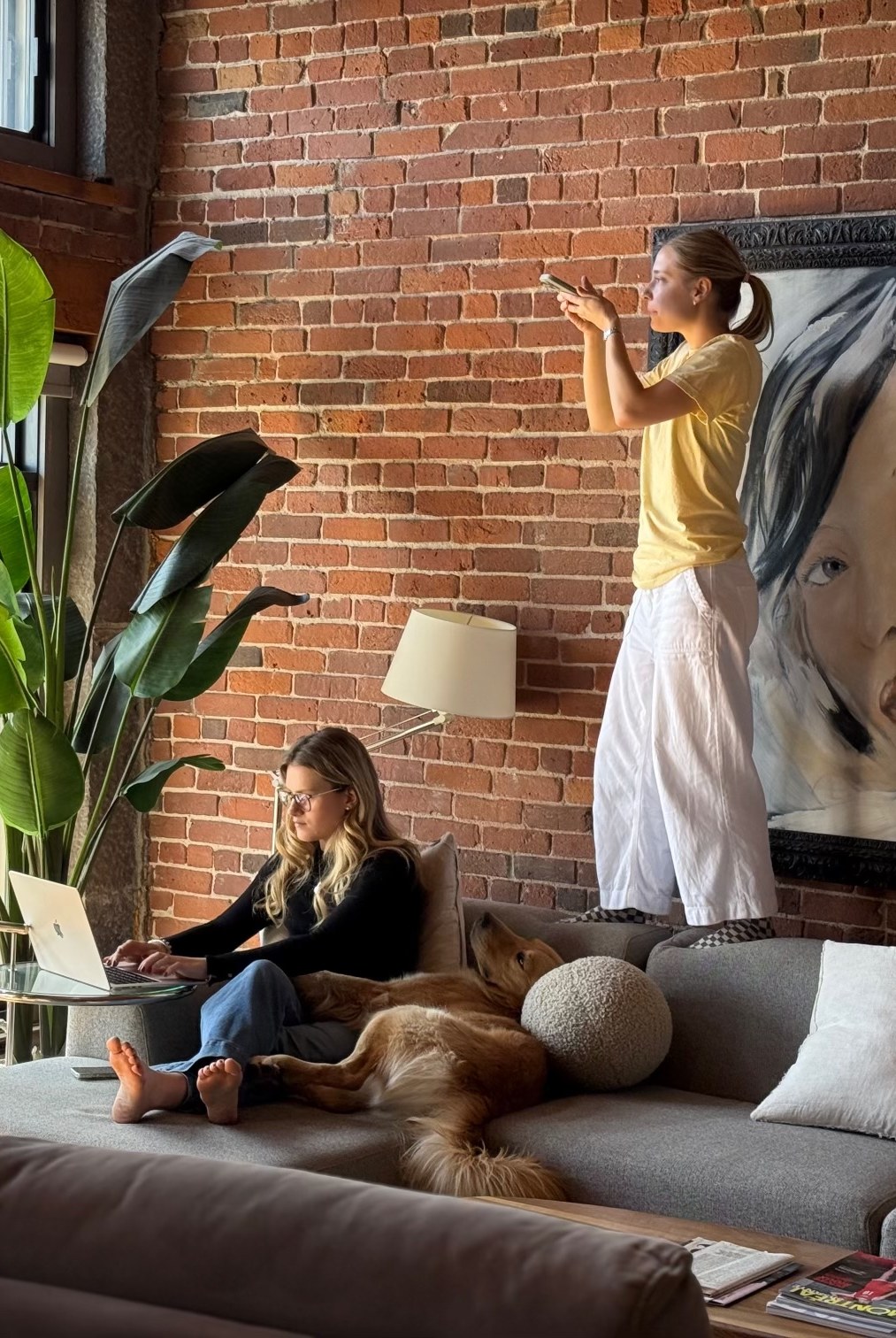 Dans un loft en brique, une femme travaille sur son portable avec un golden retriever pendant qu’une autre photographie la scène avec son téléphone.